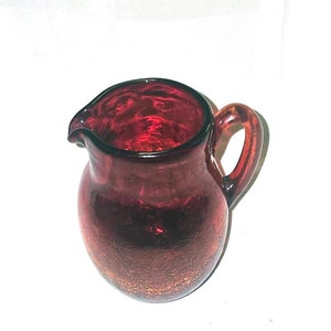 May include: A small, red glass pitcher with a textured surface and a handle. The pitcher is about 5 inches tall.