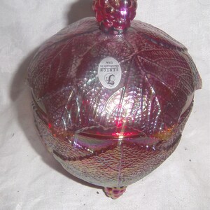 May include: A red iridescent glass bowl with a raspberry design. The bowl has a lid with a raspberry finial. The bowl is marked "FENTON Handmade in USA".