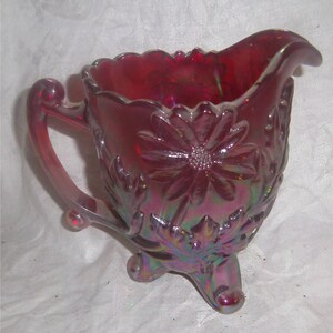 May include: A red iridescent glass creamer with a floral design and three legs. The creamer has a handle and a spout.