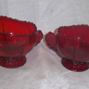 May include: A set of two red glass creamer and sugar bowls with a ruffled edge and a footed base.