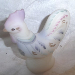 May include: A white milk glass rooster figurine with hand-painted details in shades of purple, blue, green, and pink. The rooster is standing on a white base.