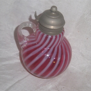 May include: A pink and white striped glass creamer with a pewter lid. The creamer has a rounded body and a handle.