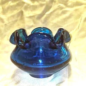 May include: A small, cobalt blue glass vase with a ruffled rim. The vase has a rounded, bulbous shape and is made of textured glass.