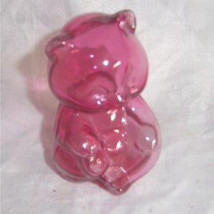 May include: A pink glass figurine of a raccoon sitting on its haunches. The figurine is made of clear glass with a pink tint.