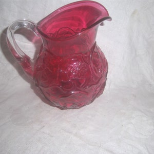 May include: A ruby red glass pitcher with a clear glass handle. The pitcher has a textured, floral design.