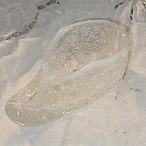 May include: A clear, glass shoe-shaped decorative piece with a detailed starburst pattern. The open-topped shoe has a rounded toe and a curved opening. The glass is transparent, allowing light to pass through, and sits on a white, textured surface.