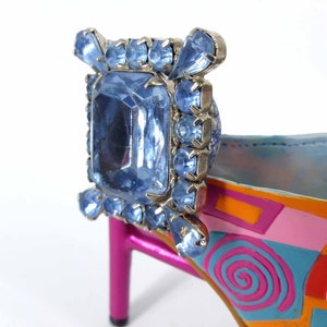 May include: A bright pink high heel shoe with a blue and orange geometric design. The shoe has a large blue rhinestone embellishment on the back.