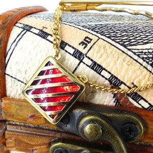 May include: A gold chain necklace with a red and gold square pendant hangs open on a wooden treasure chest with a gold clasp. The chest is decorated with a map-like design.