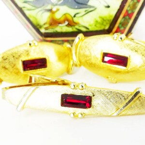 May include: A set of gold-toned cufflinks and a tie bar with red rectangular stones. The cufflinks are oval shaped and the tie bar is rectangular.