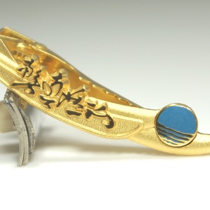 May include: A gold tie clip with a black floral design and a blue circle with a wave design. The tie clip is resting on a silver and ivory razor.