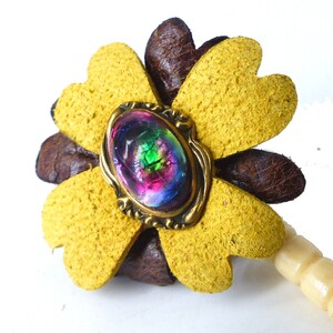 May include: A yellow leather flower with a brown leather center and a colorful iridescent glass cabochon in the center. The flower is attached to a wooden stick.