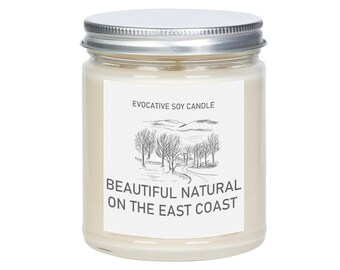 May include: A white soy candle in a glass jar with a silver lid. The label says "Evocative Soy Candle" and "Beautiful Natural on the East Coast". The label also features a black and white illustration of a winding road through a forest.