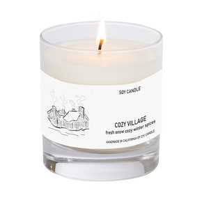 K&ouml;nnte beinhalten: Eine wei&szlig;e Sojawachskerze in einem durchsichtigen Glasgef&auml;&szlig; mit einer Schwarzwei&szlig;illustration eines Hauses mit Rauch aus dem Schornstein. Das Etikett lautet "SOY CANDLE", "COZY VILLAGE", "fresh snow cozy winter spices" und "HANDMADE IN CALIFORNIA BY CHI CANDLE".