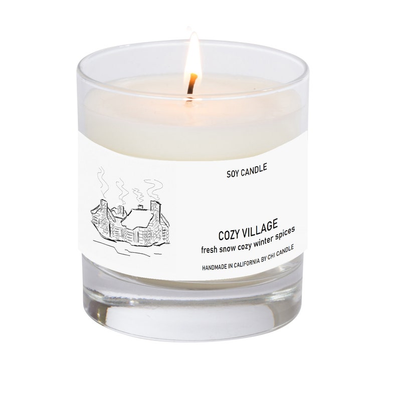 K&ouml;nnte beinhalten: Eine wei&szlig;e Sojawachskerze in einem durchsichtigen Glasgef&auml;&szlig; mit einer Schwarzwei&szlig;illustration eines Hauses mit Rauch aus dem Schornstein. Das Etikett lautet "SOY CANDLE", "COZY VILLAGE", "fresh snow cozy winter spices" und "HANDMADE IN CALIFORNIA BY CHI CANDLE".