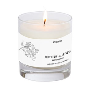 May include: A white soy candle in a clear glass jar with a black and white label that reads "Protection + Alertness eucalyptus mint Handmade in California by Chi Candle" and a drawing of a sprig of eucalyptus.