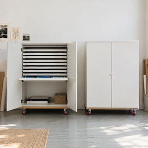 May include: Two white storage cabinets on casters, each featuring double doors. The left cabinet is open, revealing multiple horizontal sliding shelves for flat storage and a lower compartment containing books and a box. The right cabinet is closed.