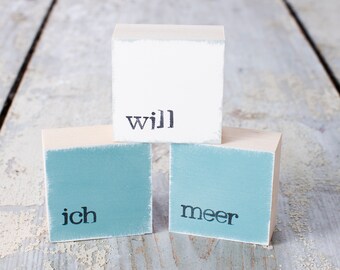 May include: Three wooden blocks stacked on top of each other. The top block is white with the word "will" printed in black. The bottom two blocks are light blue with the words "ich" and "meer" printed in black.