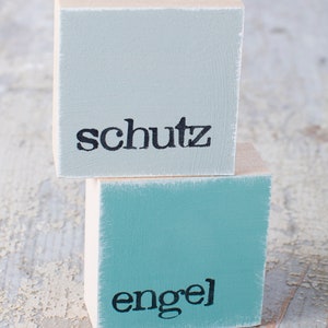 May include: Two wooden blocks stacked on top of each other. The top block is light blue and says "schutz" in black lettering. The bottom block is teal blue and says "engel" in black lettering.