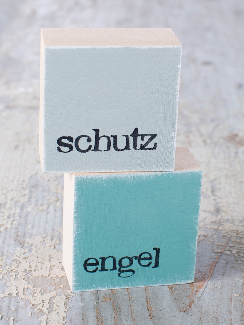 May include: Two wooden blocks stacked on top of each other. The top block is light blue and says "schutz" in black lettering. The bottom block is teal blue and says "engel" in black lettering.