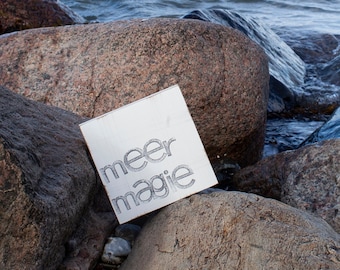 May include: A white wooden sign with the text "meer magie" written in black ink. The sign is resting on a large, gray rock.