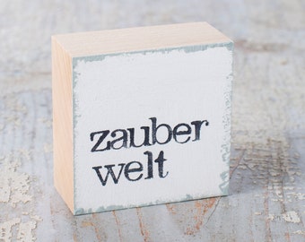 May include: A small wooden block with a white painted surface and the text "zauber welt" in black lettering.