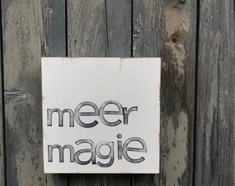 May include: White wooden sign with black text that reads "meer magie" against a weathered gray wooden background.