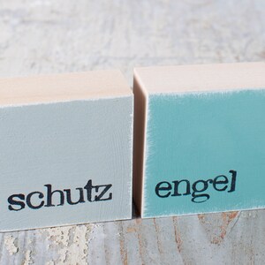 May include: Two wooden blocks painted in light blue and turquoise with the words "schutz" and "engel" painted in black.