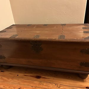 May include: A vintage wooden chest, rectangular in shape, crafted from dark brown wood. It features metal corner and edge reinforcements. The chest stands on short, rounded feet and has a decorative metal lock plate.