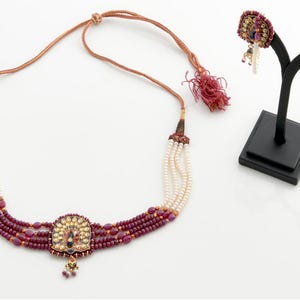 Peut inclure: Un ensemble de collier et de boucles d'oreilles orné. Le collier présente plusieurs rangées de perles blanches et de perles bordeaux, avec un pendentif central. Les boucles d'oreilles sont présentées sur un présentoir noir. Les bijoux ont des accents dorés et de petits détails colorés.