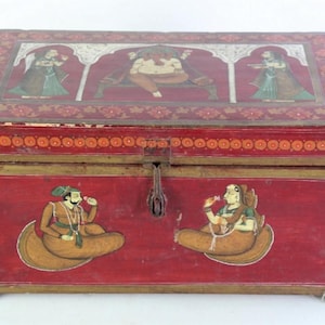 Puede incluir: Una caja de madera roja con intrincados diseños pintados en oro y blanco. La caja presenta escenas de personas y un elefante, todos pintados en un estilo tradicional. La caja tiene una tapa con bisagras y un cierre de metal.