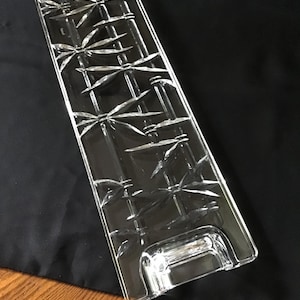 May include: A clear, rectangular glass serving tray featuring an embossed bamboo stalk and leaf pattern across its surface. The tray has integrated handles at both ends, with a smooth, polished finish that catches the light, creating a reflective effect.