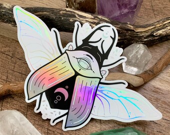 Holographic vinyl sticker "Luna Beetle" picturing magical horned eyed beetle decorated with crystals, herbs and moon phases. 5.9"W x 3.8"H.