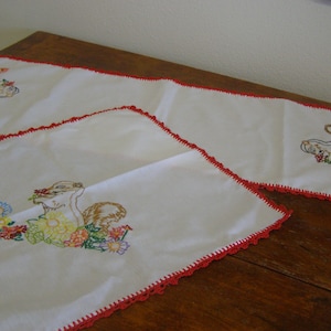 May include: Two white embroidered table runners with red crocheted edges. One runner features a squirrel with flowers and the other features a girl with flowers.