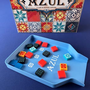Op de afbeelding: Het bordspel "Azul" wordt getoond, met een doos met kleurrijke tegelpatronen en de titel van het spel. Een blauwe tray bevat spelstukken: rode, zwarte, gele en blauwe vierkante tegels met decoratieve patronen. Het spel staat tegen een blauwe achtergrond.