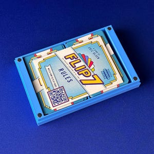Puede incluir: Una caja azul rectangular que contiene una baraja de cartas. Las cartas est&aacute;n etiquetadas como "FLIP 7 RULES" con un c&oacute;digo QR y un dise&ntilde;o colorido. El juego es de Eric Olsen.