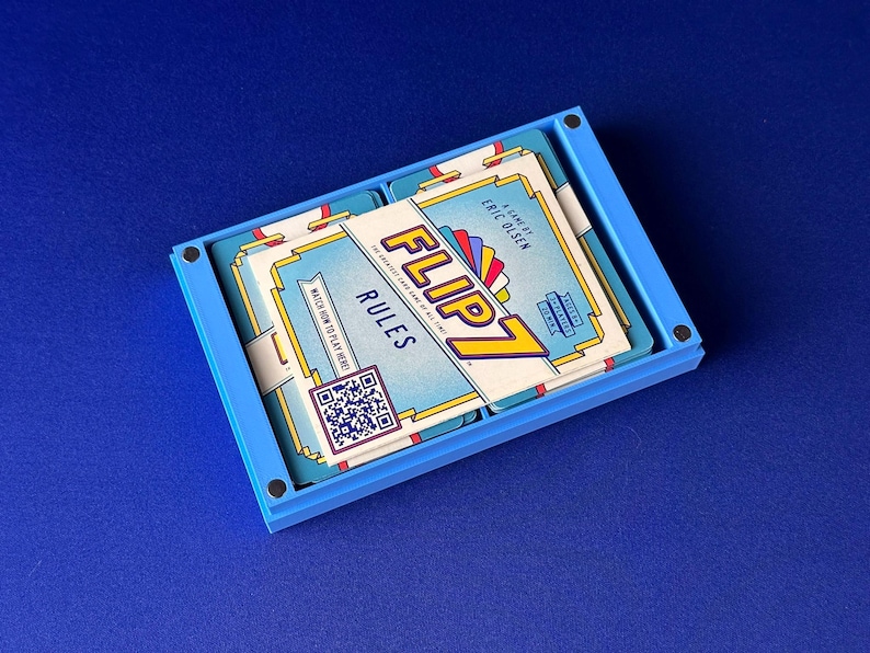 Puede incluir: Una caja azul rectangular que contiene una baraja de cartas. Las cartas est&aacute;n etiquetadas como "FLIP 7 RULES" con un c&oacute;digo QR y un dise&ntilde;o colorido. El juego es de Eric Olsen.