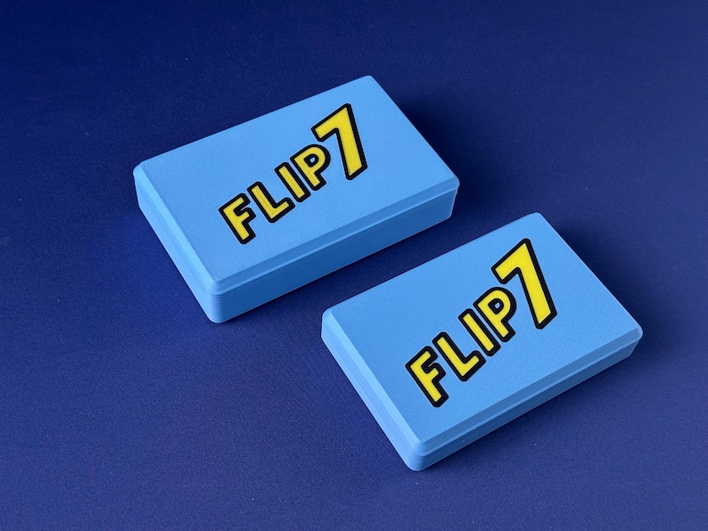Puede incluir: Dos cajas rectangulares de color azul claro con el texto "FLIP 7" en amarillo con un contorno negro. Las cajas est&aacute;n sobre una superficie azul oscuro. Las cajas parecen ser cajas de juego.