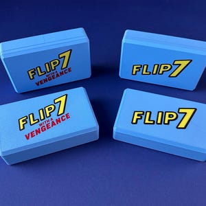 Puede incluir: Cuatro cajas de juego azul claro con el texto "FLIP 7" en amarillo y negro, y "WITH A VENGEANCE" en rojo. Las cajas rectangulares tienen una superficie ligeramente texturizada, sobre un fondo azul oscuro.