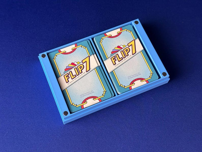 Puede incluir: Un soporte de cartas azul claro que contiene dos barajas de cartas Flip 7. Las cartas tienen un fondo azul claro con el t&iacute;tulo del juego en una pancarta blanca. El soporte de cartas est&aacute; sobre una superficie azul.