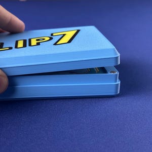 Puede incluir: Una caja de juego azul claro con el texto "FLIP 7" en amarillo y negro. La caja est&aacute; parcialmente abierta, revelando las cartas del juego en su interior. La caja est&aacute; sobre una superficie azul.