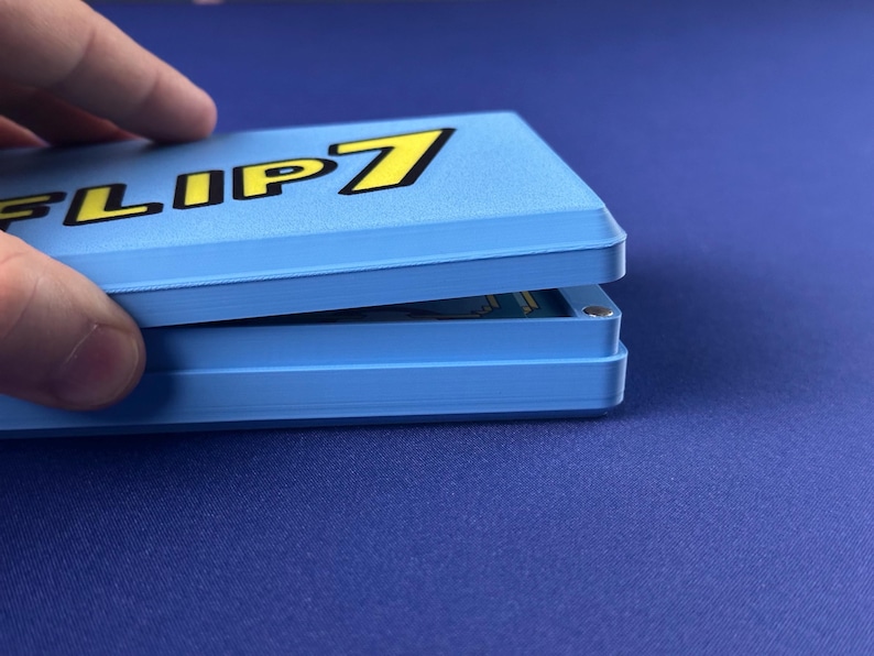 Puede incluir: Una caja de juego azul claro con el texto "FLIP 7" en amarillo y negro. La caja est&aacute; parcialmente abierta, revelando las cartas del juego en su interior. La caja est&aacute; sobre una superficie azul.