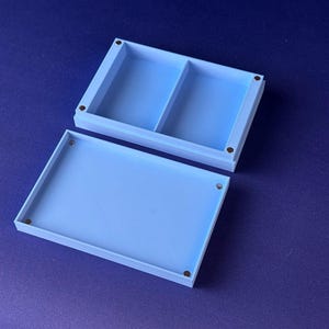 Puede incluir: Una caja rectangular azul claro con tapa, abierta hacia un lado. La caja est&aacute; dividida en dos compartimentos. Se ven peque&ntilde;os imanes circulares de color oscuro en cada esquina de la caja y la tapa. El fondo es azul oscuro liso.