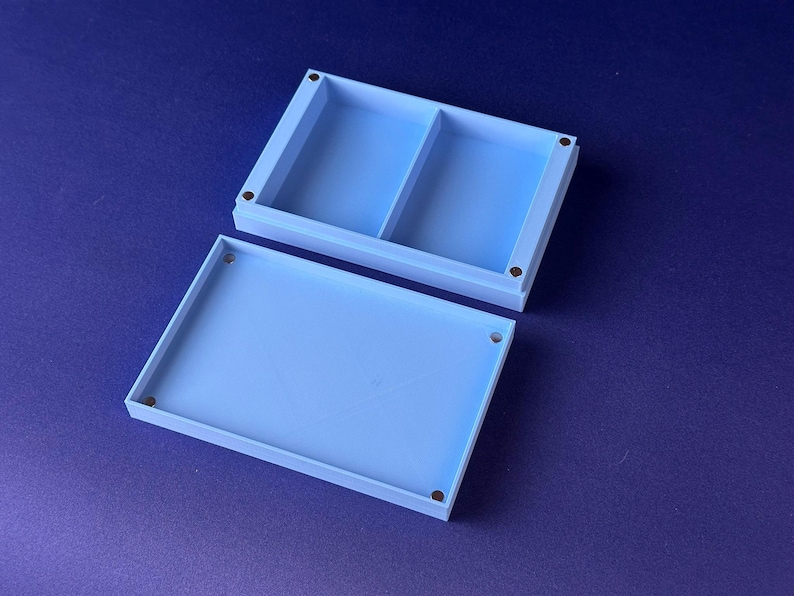 Puede incluir: Una caja rectangular azul claro con tapa, abierta hacia un lado. La caja est&aacute; dividida en dos compartimentos. Se ven peque&ntilde;os imanes circulares de color oscuro en cada esquina de la caja y la tapa. El fondo es azul oscuro liso.