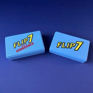 Puede incluir: Dos cajas rectangulares azul claro con el texto "FLIP 7" en amarillo y negro, y "WITH A VENGEANCE" en rojo. Las cajas est&aacute;n sobre una superficie azul oscuro.