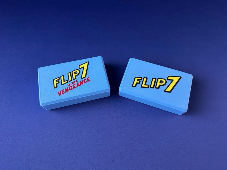 Puede incluir: Dos cajas rectangulares azul claro con el texto "FLIP 7" en amarillo y negro, y "WITH A VENGEANCE" en rojo. Las cajas est&aacute;n sobre una superficie azul oscuro.