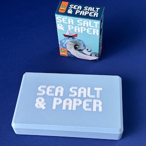 Könnte beinhalten: Eine rechteckige hellblaue Aufbewahrungsbox mit dem in Weiß aufgedruckten Text SEA SALT & PAPER auf dem Deckel, begleitet von einer vertikalen Spieleschachtel mit demselben Titel und einer Abbildung eines Origami-Boots auf einer Welle.