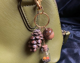 May include: A gold-colored keychain with a pinecone, acorn, and tiny glass bottle charm. The pinecone is brown, the acorn is brown and tan, and the bottle contains small orange and yellow objects. The keychain is attached to a green handbag.