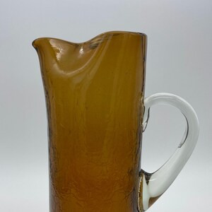 Puede incluir: Jarra de vidrio color ámbar con superficie texturizada y asa curva transparente. La jarra tiene forma alta y cilíndrica y un pico vertedor. Un artículo decorativo y funcional para servir bebidas.