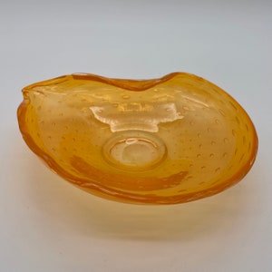 May include: An amber-coloured glass bowl with an unusual, irregular shape. The bowl has small, bubble-like inclusions throughout the glass, adding texture. The rim of the bowl has a wavy design. The bowl is likely for decorative purposes.