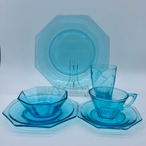 Può includere: Un set di vetreria azzurra, tra cui un grande piatto ottagonale, un bicchiere, una tazza da tè con piattino e una piccola ciotola con piattino. Il set è realizzato in vetro trasparente ed è disposto su una superficie bianca.